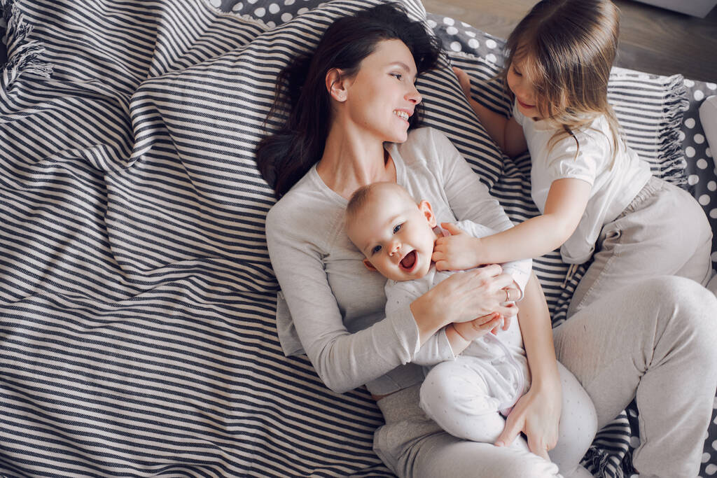 Egy fiatal mosolygó anya játszik két kislánnyal egy csíkos ágyon. A gyerekek nevetnek. - Fotó, kép