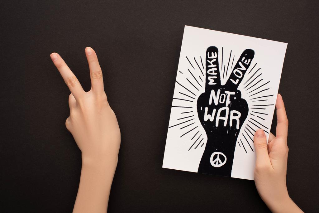 обрізаний вид жінки, що демонструє знак миру і тримає білий папір з малюнком рук і робить любов не війною на чорному тлі
 - Фото, зображення