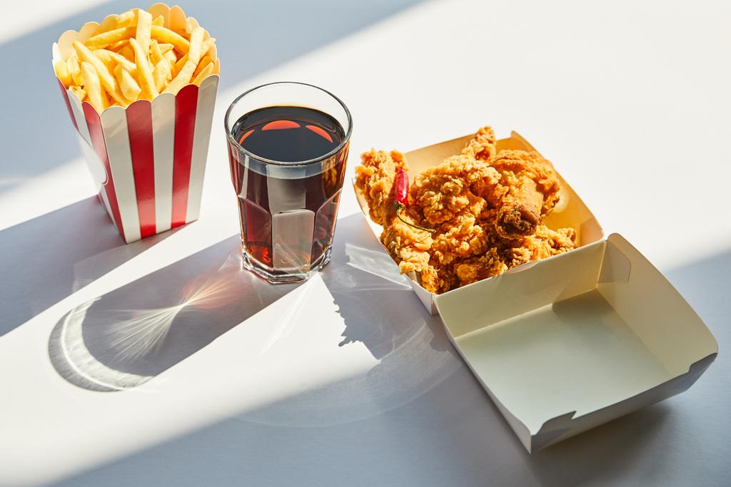 smaczny smażony kurczak, frytki i soda w szkle na białym stole w słońcu - Zdjęcie, obraz