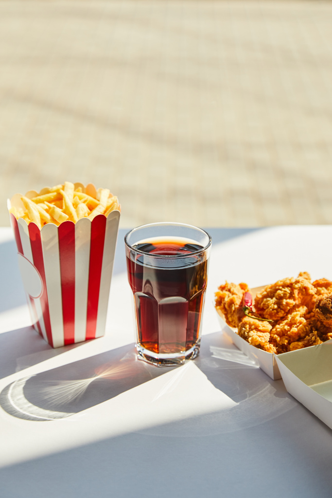 gustoso pollo fritto, patatine fritte e soda in vetro su tavolo bianco alla luce del sole vicino alla finestra
 - Foto, immagini