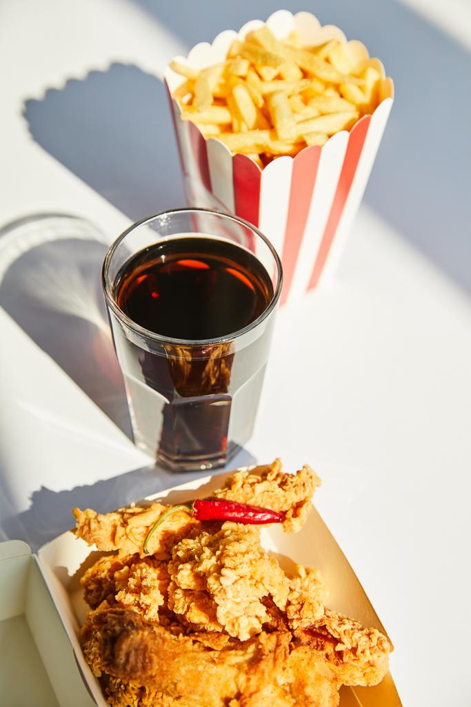 selektywne skupienie smacznego smażonego kurczaka, frytek i sody w szkle na białym stole w słońcu - Zdjęcie, obraz