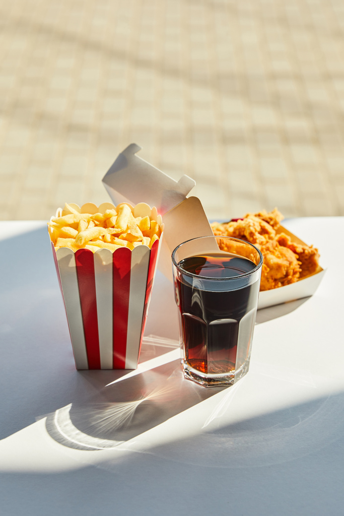 窓の近くの太陽の下で白いテーブルの上においしい揚げ鶏、フランスのフライドポテトとソーダの選択的な焦点 - 写真・画像