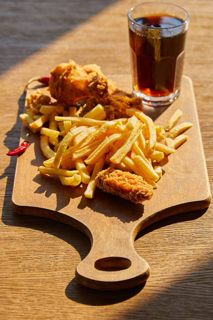 enfoque selectivo de pollo frito picante, papas fritas a bordo con soda en vidrio sobre mesa de madera a la luz del sol
 - Foto, imagen