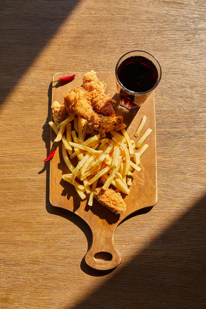vista superior de pollo frito picante, papas fritas a bordo con soda en vidrio sobre mesa de madera a la luz del sol
 - Foto, Imagen