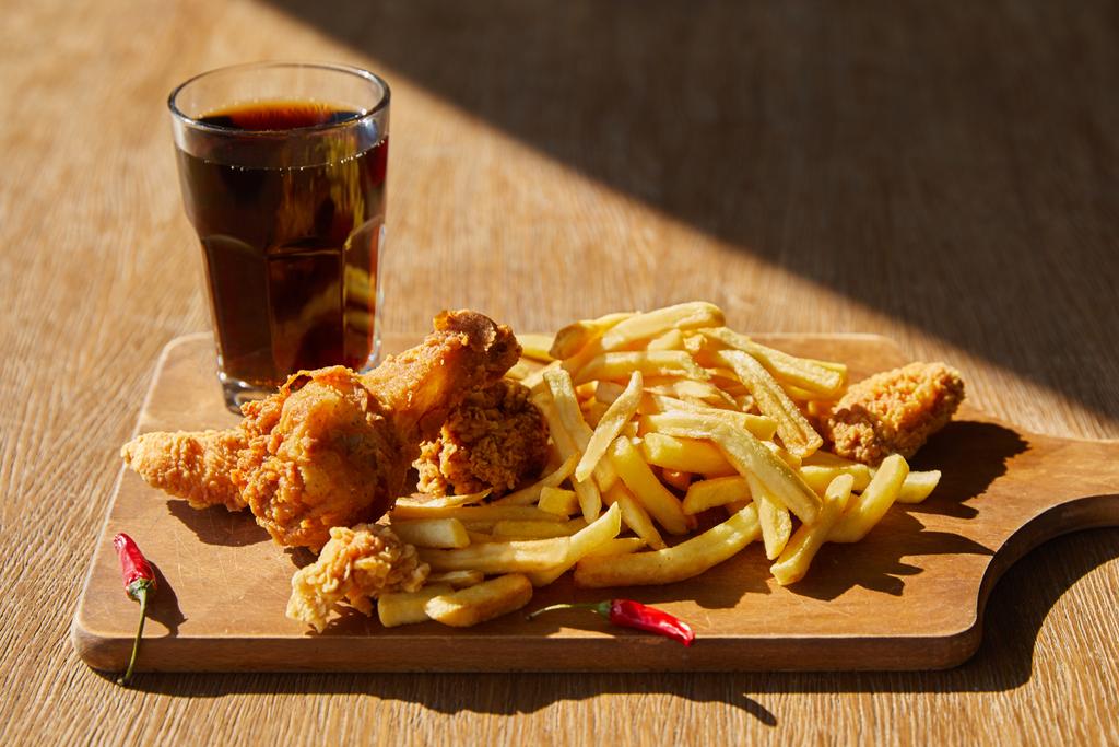 spicy deep fried chicken, french fries on board with soda in glass on wooden table in sunlight - Photo, Image