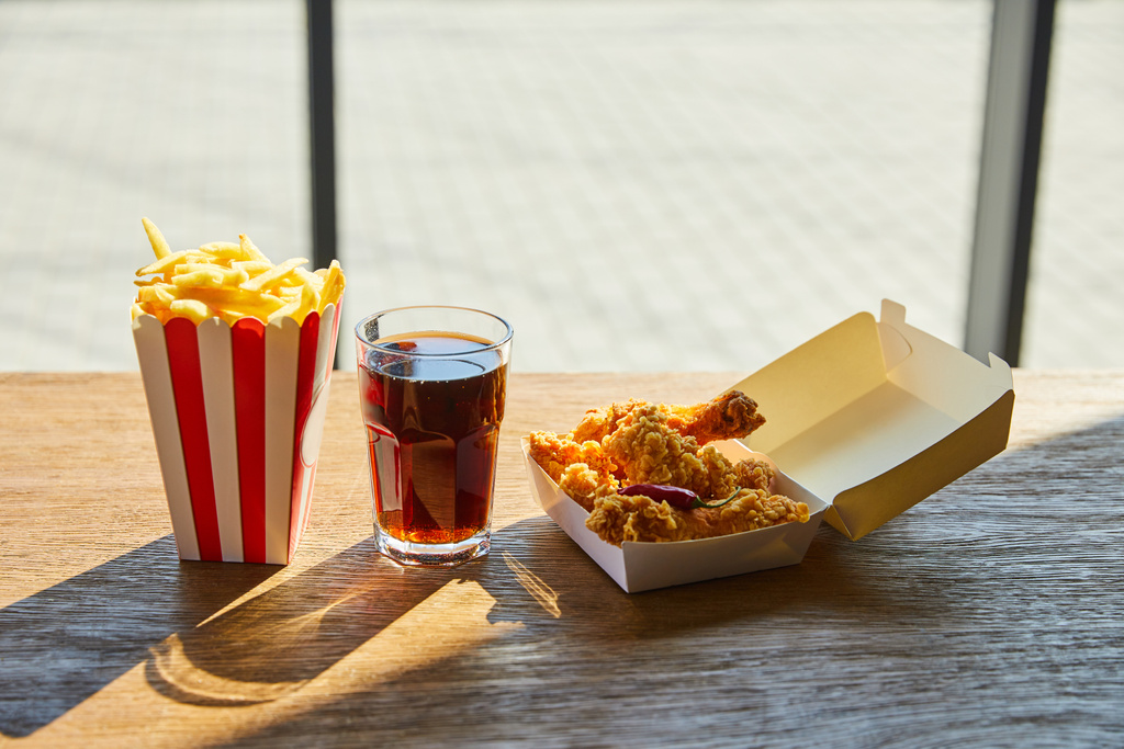 pikantny smażony kurczak, frytki i soda w szkle na drewnianym stole w słońcu przy oknie - Zdjęcie, obraz