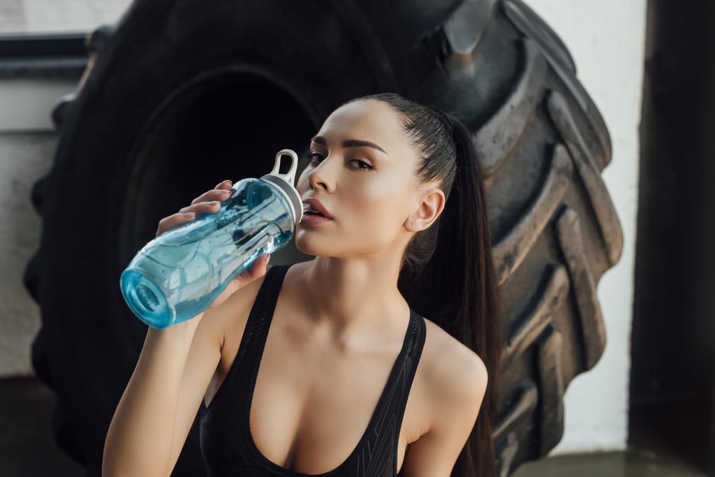 Σέξι αθλήτρια πίνει νερό και κοιτάζει την κάμερα κοντά στο λάστιχο στο γυμναστήριο - Φωτογραφία, εικόνα
