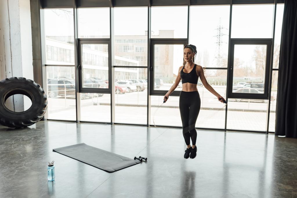 Женщина прыгает с трамплина рядом с фитнес-ковриком, гантелями и спортивной бутылкой в спортзале
 - Фото, изображение
