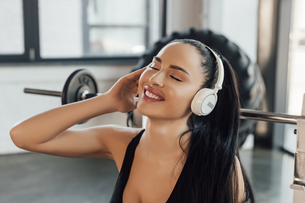 Krásná sportovkyně se zavřenýma očima s úsměvem a posloucháním hudby ve sluchátkách v tělocvičně - Fotografie, Obrázek