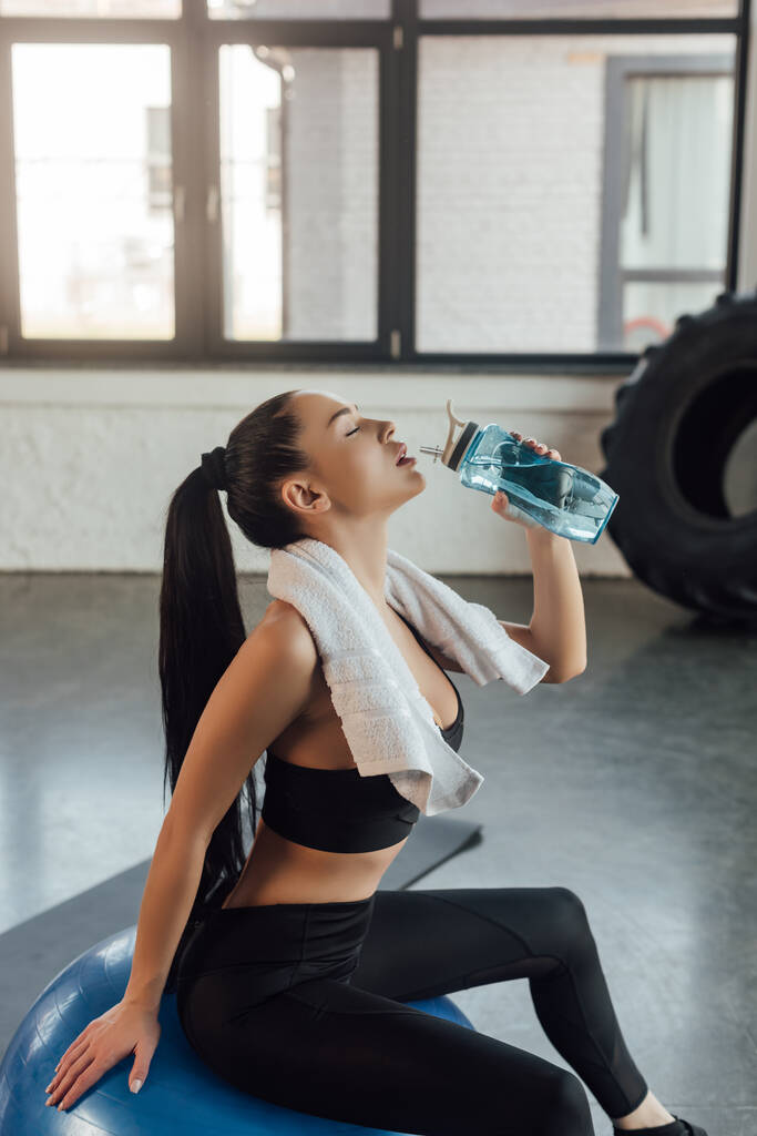 Όμορφη αθλήτρια με κλειστά μάτια και πετσέτα πόσιμο νερό και κάθεται σε μπάλα γυμναστικής στο γυμναστήριο - Φωτογραφία, εικόνα