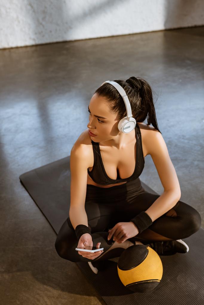 Hochwinkelaufnahme einer Sportlerin mit überkreuzten Beinen in Kopfhörern, die ihr Smartphone in Ballnähe auf der Fitnessmatte im Fitnessstudio hält - Foto, Bild