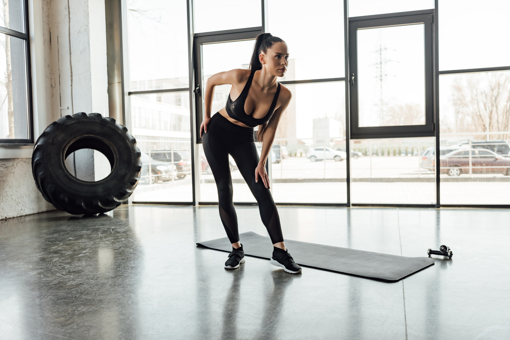Barna sportos nő hajlik közelében fitness mat és súlyzók tornaterem - Fotó, kép