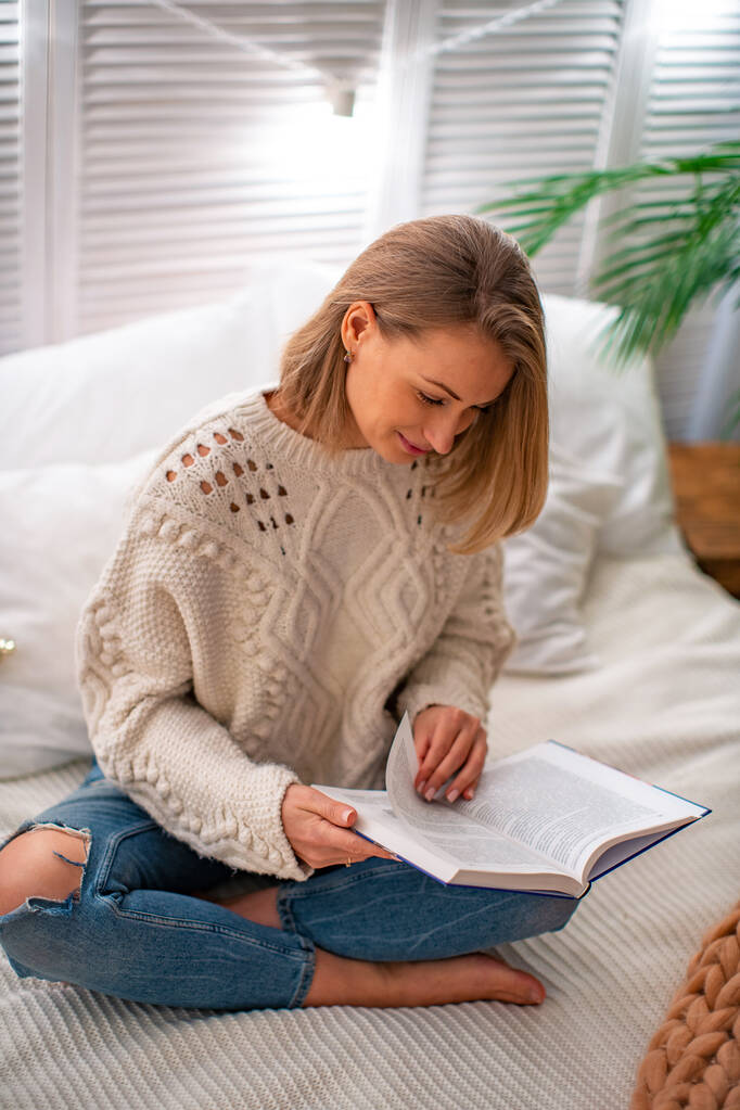 fiatal mosolygós nő ül az ágyon egy könyvvel. A szőke az oktatási irodalmat tanulmányozza. Az esti olvasmány. A szabadidő fogalma. Maradj otthon. - Fotó, kép