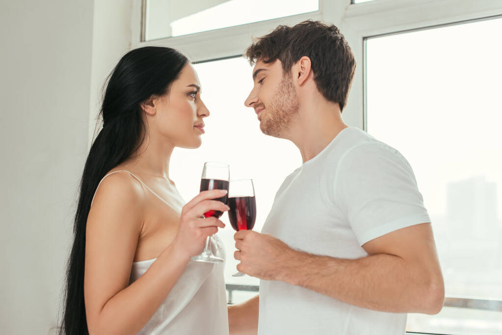 όμορφο ζευγάρι κρατώντας ποτήρια με κόκκινο κρασί και κοιτάζοντας ο ένας τον άλλο κοντά στο παράθυρο κατά τη διάρκεια της αυτοαπομόνωσης  - Φωτογραφία, εικόνα