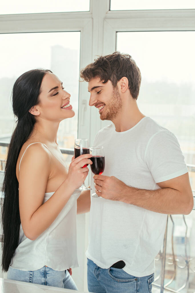 belo casal sorridente segurando copos com vinho tinto durante o auto-isolamento perto da janela
  - Foto, Imagem