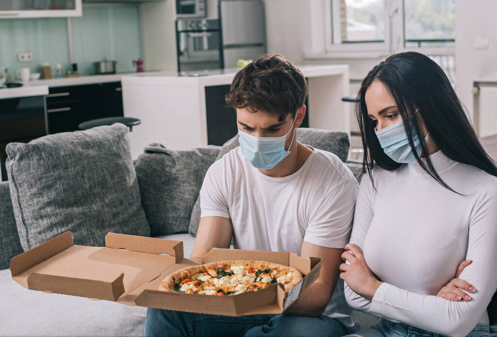 ziek paar in medische maskers kijken naar pizza thuis tijdens zelfisolatie - Foto, afbeelding