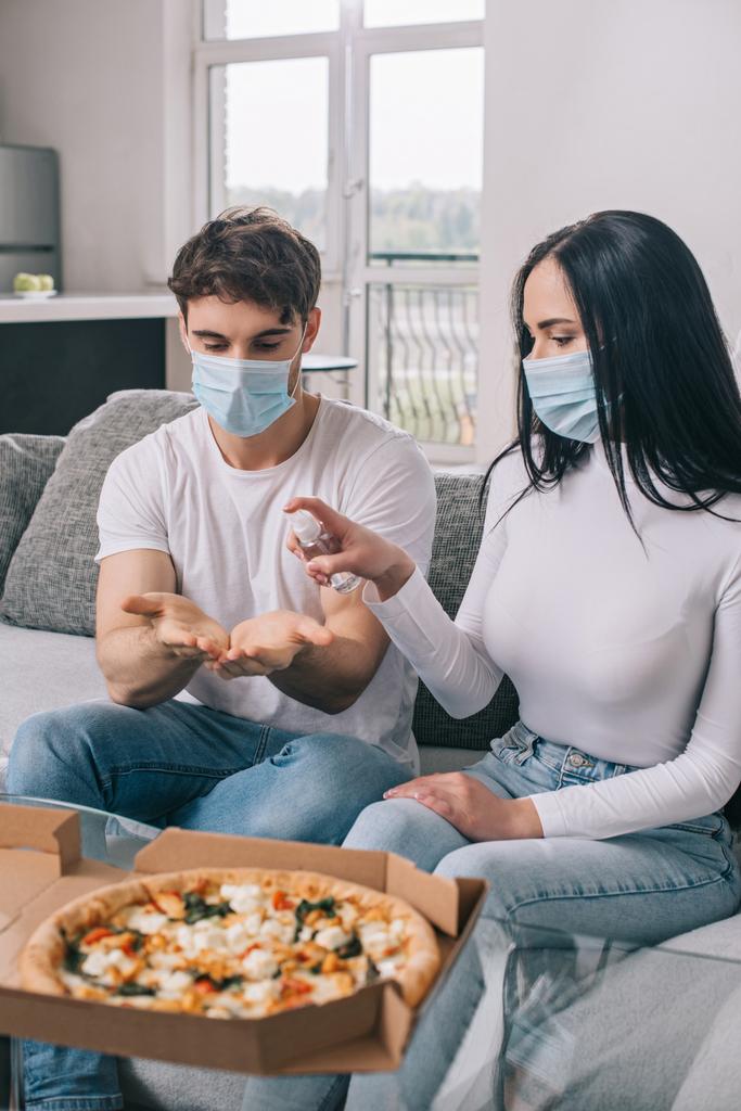 pareja enferma en máscaras médicas usando aerosol antiséptico antes de comer pizza en casa durante el auto aislamiento
 - Foto, Imagen