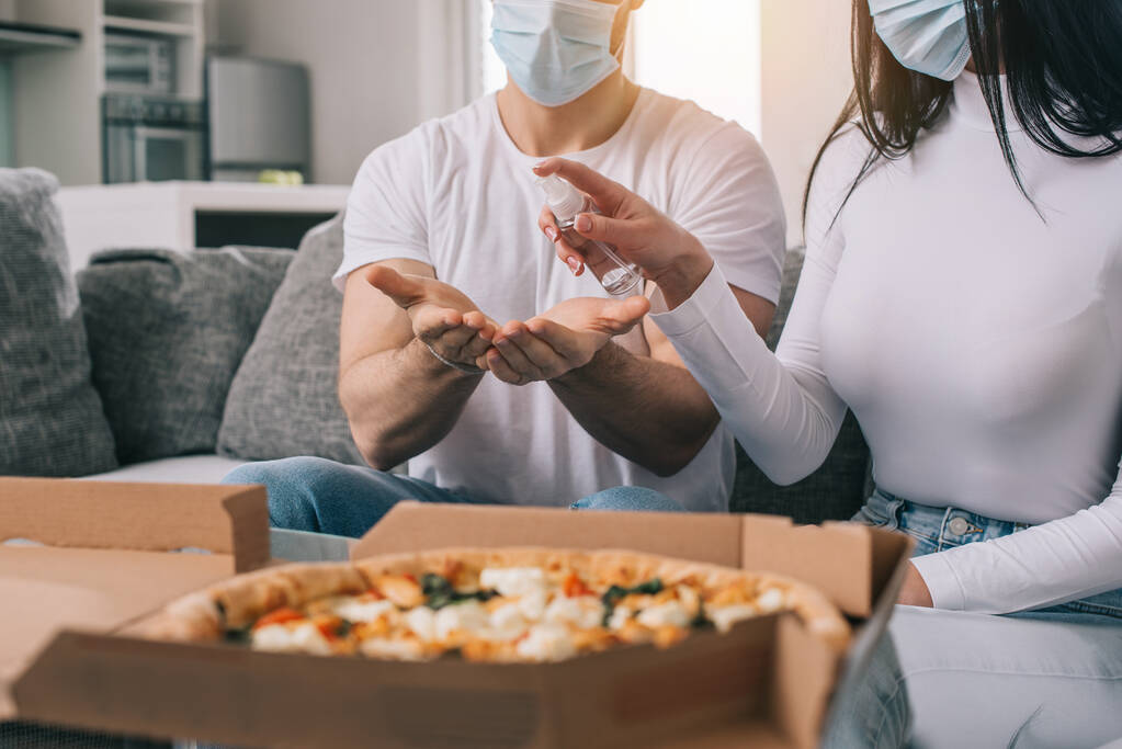 обрезанный вид больной пары в медицинских масках с помощью антисептического спрея перед употреблением пиццы в домашних условиях во время самоизоляции
 - Фото, изображение