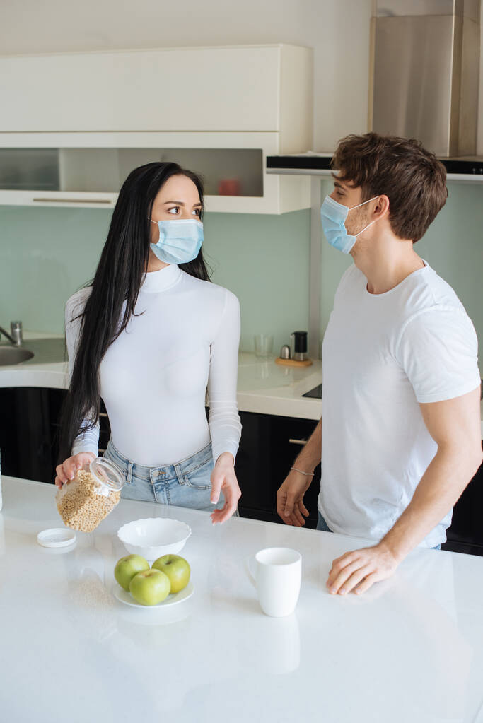 jovem casal em máscaras médicas tomando café da manhã em casa durante o auto-isolamento
 - Foto, Imagem