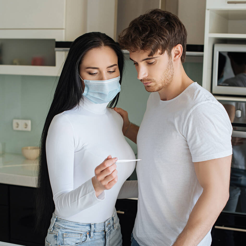 boyfriend and ill girl in medical mask looking at thermometer during self isolation - Photo, Image