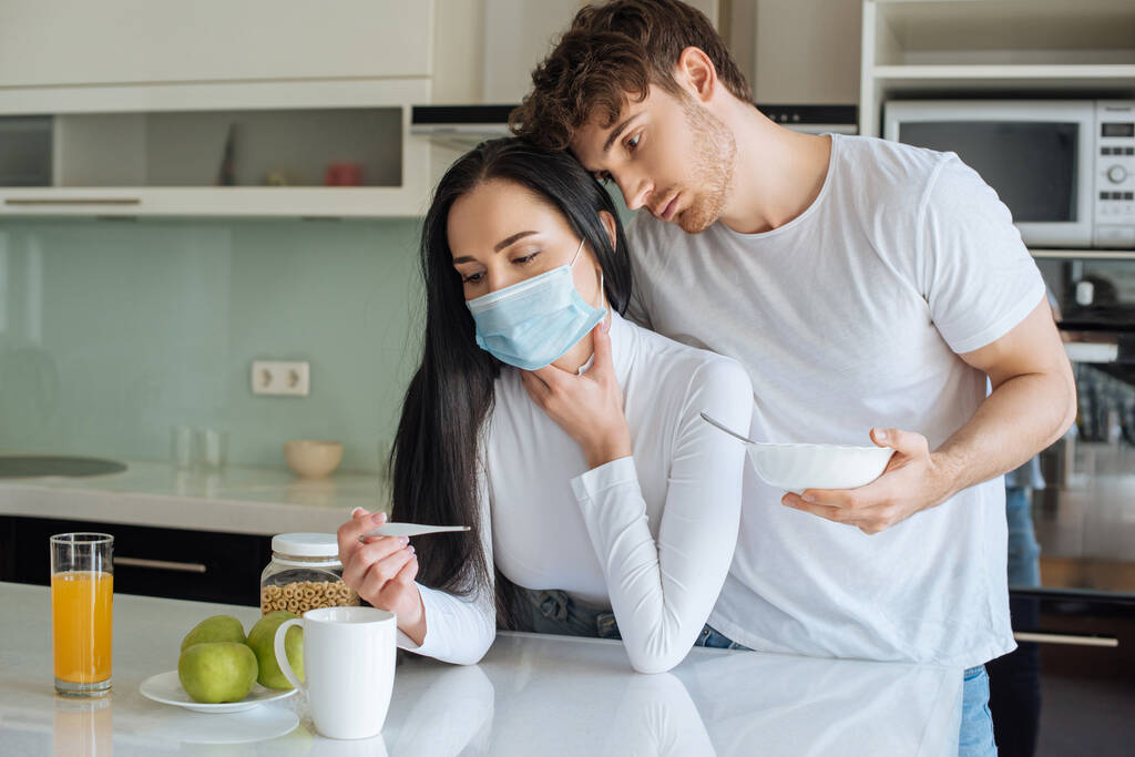 férfi és beteg nő orvosi maszkban nézi a hőmérőt és reggelizik az elszigeteltség alatt - Fotó, kép