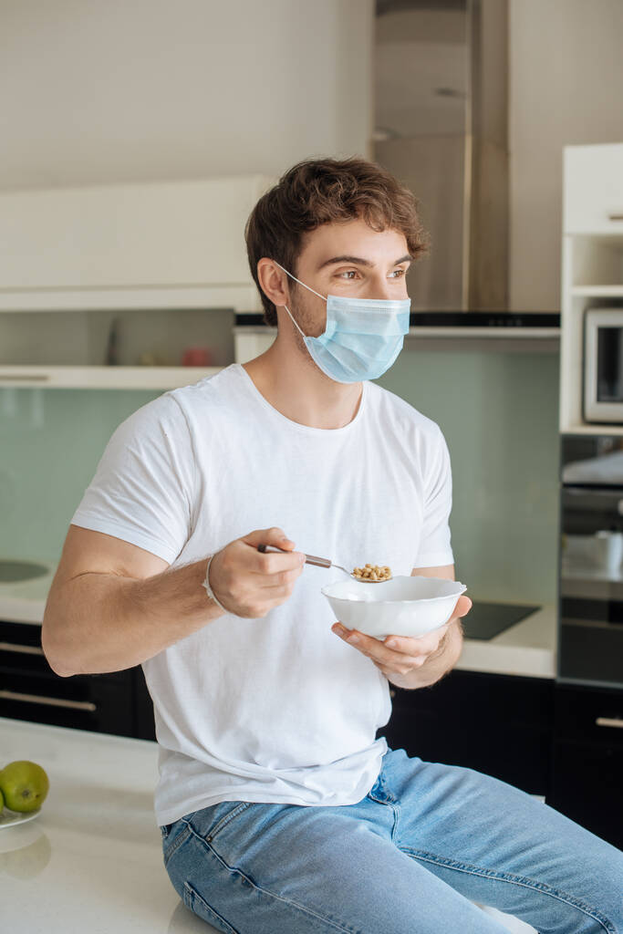 όμορφος άρρωστος άνδρας με ιατρική μάσκα κρατώντας μπολ με κορν φλέικς στην κουζίνα σε αυτο-απομόνωση  - Φωτογραφία, εικόνα