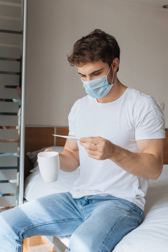 trauriger kranker Mann in medizinischer Maske hält Thermometer und Tasse mit Heißgetränk während der Selbstisolierung auf dem Bett - Foto, Bild