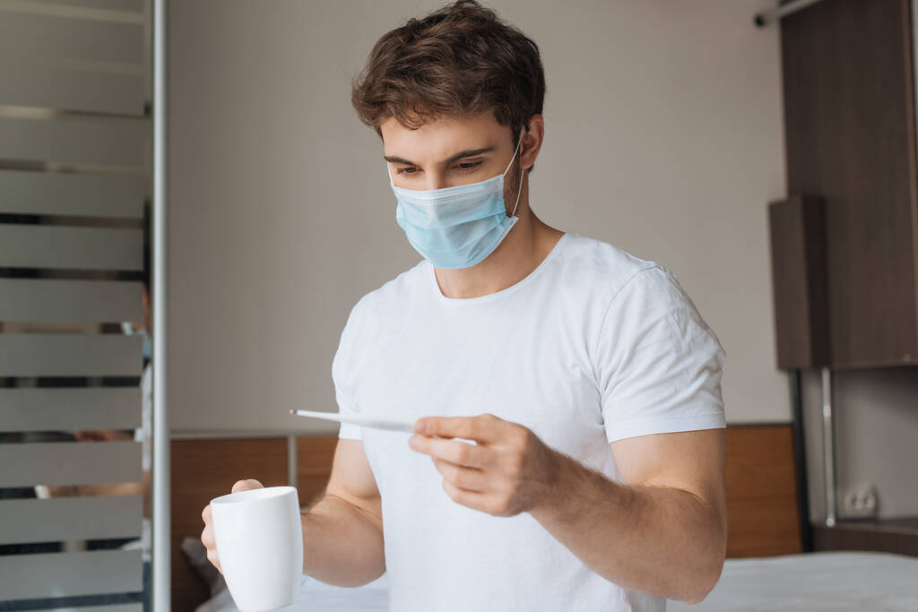 hombre enfermo con máscara médica sosteniendo termómetro y taza con bebida caliente durante el auto aislamiento
 - Foto, Imagen