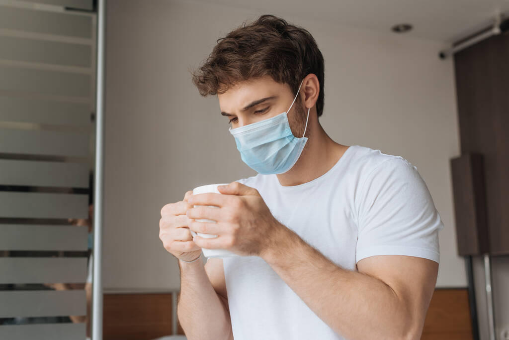 Tıbbi maskeli hasta bir adam kendini izole ederken yatak odasında sıcak içecekle fincana bakıyor. - Fotoğraf, Görsel