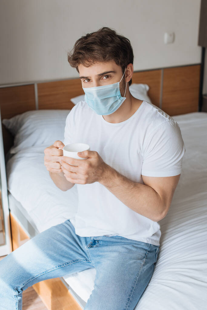 κουρασμένος άρρωστος άνθρωπος σε ιατρική μάσκα κρατώντας κύπελλο με ζεστό ρόφημα στο κρεβάτι κατά τη διάρκεια της αυτο-απομόνωσης - Φωτογραφία, εικόνα