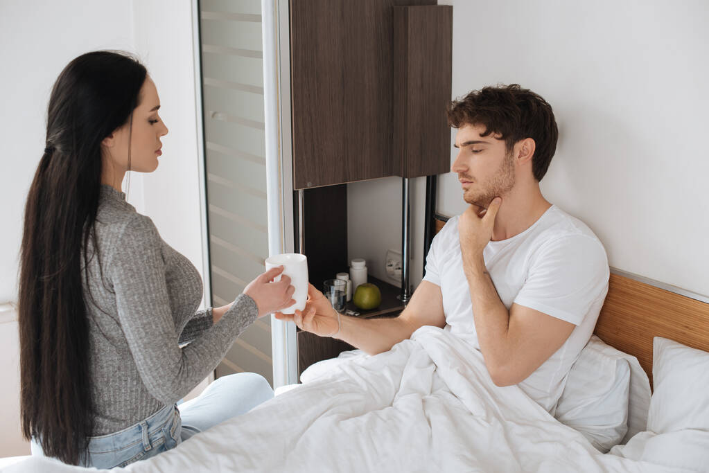 жінка дає чашку хворому чоловікові з болем у ліжку під час самоізоляції
  - Фото, зображення