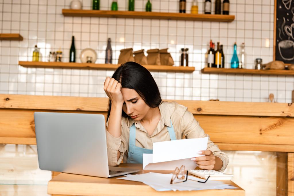 Расстроенный владелец кафе держит бумагу рядом с калькулятором, ноутбук и документы с очками за столом
 - Фото, изображение