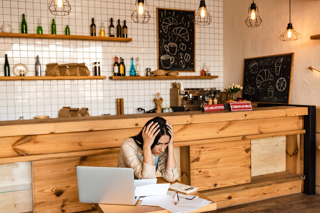 Aufgebrachter Café-Besitzer berührt Kopf in der Nähe von Laptop, Dokumenten und Taschenrechner am Tisch - Foto, Bild