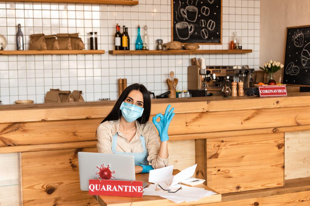 Ιδιοκτήτης καφετέριας με ιατρική μάσκα κοιτάζει την κάμερα και δείχνει εντάξει πινακίδα κοντά σε χαρτιά, laptop, γυαλιά και κάρτα με γράμματα καραντίνας στο τραπέζι - Φωτογραφία, εικόνα