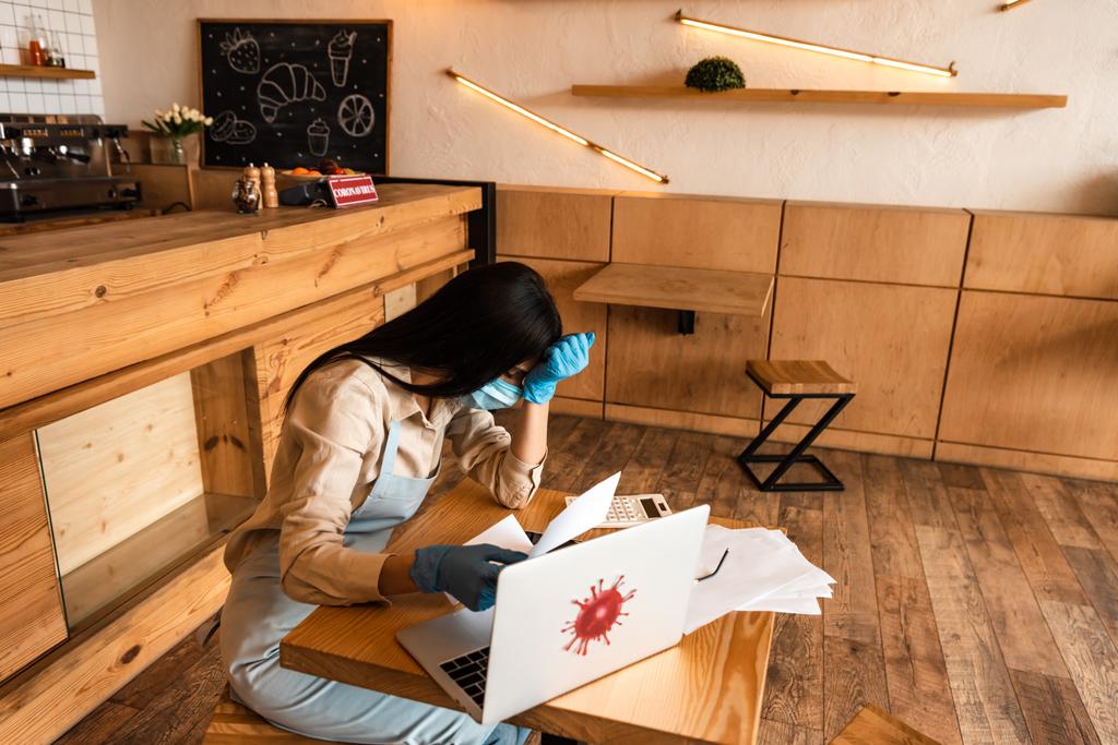 Stressed kavárna majitel v lékařské masce drží papír v blízkosti notebooku a kalkulačka u stolu - Fotografie, Obrázek