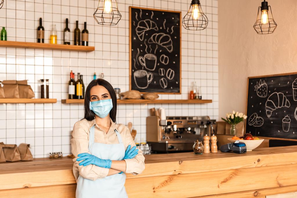 Café proprietário com braços cruzados em máscara médica e luvas de látex olhando para a câmera perto da mesa
 - Foto, Imagem