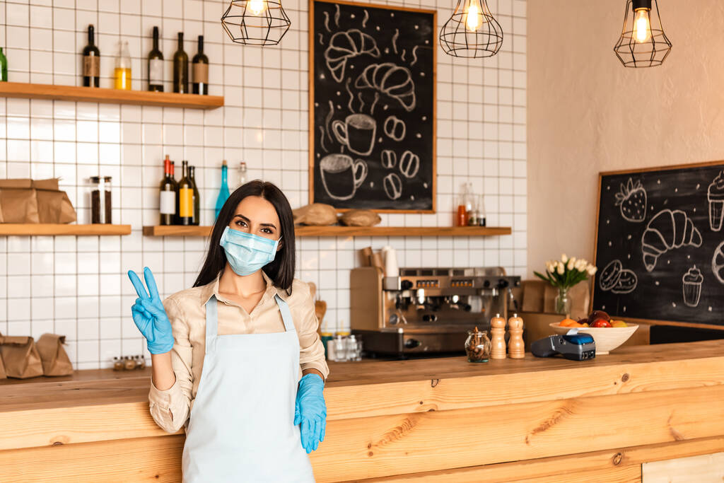 Cafe owner in medical mask and latex gloves looking at camera and showing victory sign near table - Photo, Image