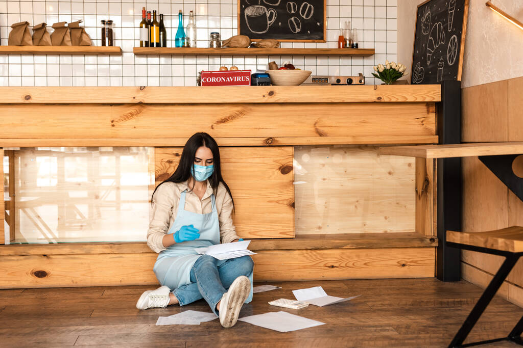 Cafébesitzer in medizinischer Maske liest Dokumente neben Tisch auf dem Fußboden - Foto, Bild