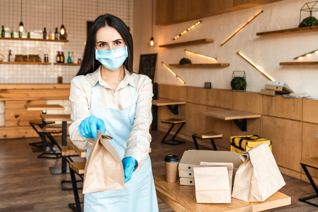 Cafe owner in medical mask showing paper bag and looking at camera near table with disposable cup of coffee and boxes - Photo, Image