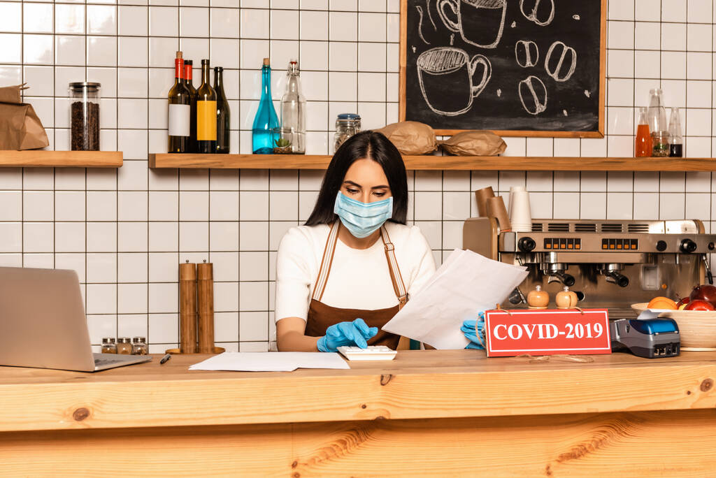 Proprietário do café em máscara médica com papéis usando calculadora perto de laptop e cartão com letras covid-2019 na mesa
 - Foto, Imagem