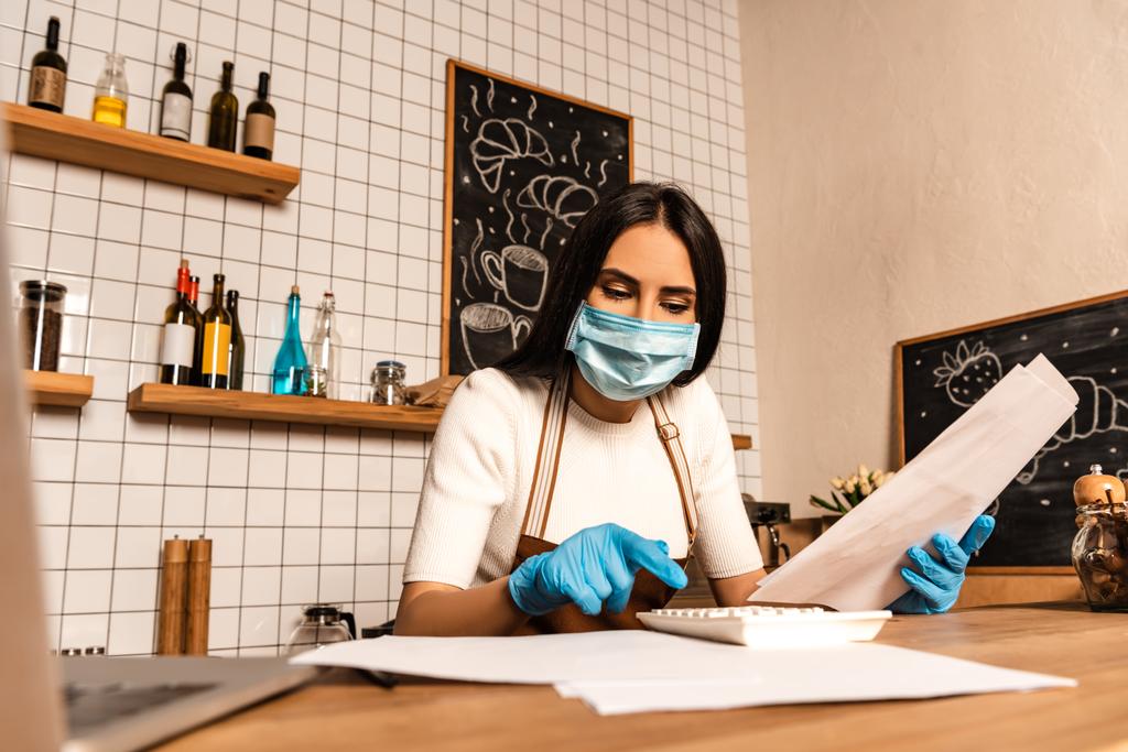 Владелец концентрированного кафе в медицинской маске с бумагами на калькуляторе возле ноутбука за столом
 - Фото, изображение