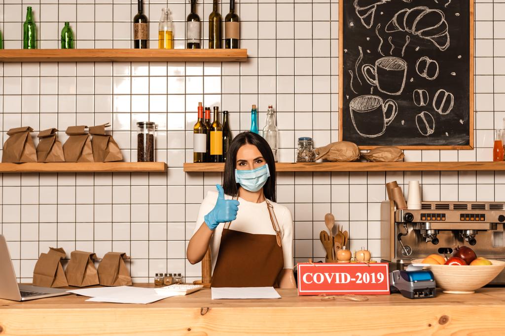 Majitel kavárny v lékařské masce ukazující jako ceduli u stolu s papíry, kartičku s nápisem covid-2019, platební terminál a mísu s ovocem - Fotografie, Obrázek