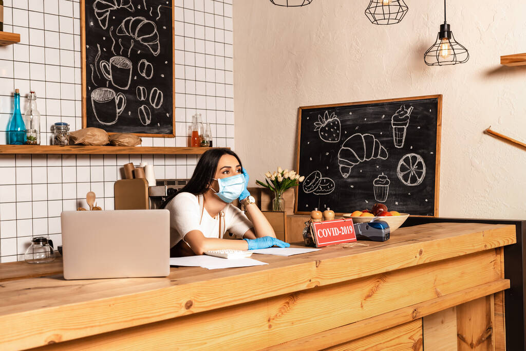 Задумчивый владелец кафе возле стола с ноутбуком, бумагами, карточкой с надписью "ковид-2019", платежным терминалом и миской с фруктами
  - Фото, изображение