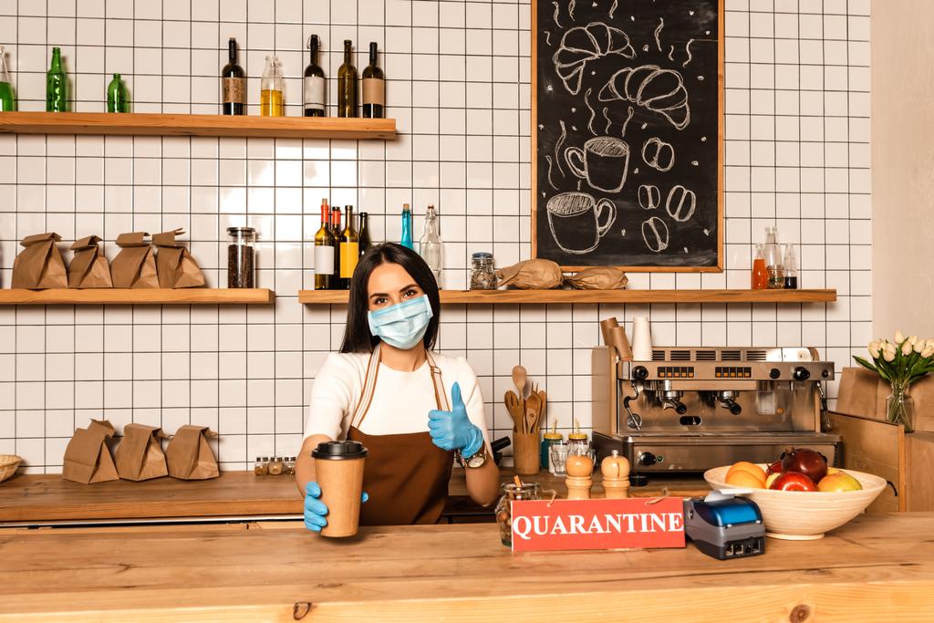 Καφενείο ιδιοκτήτης σε ιατρική μάσκα δείχνει σαν σημάδι και χάρτινο φλιτζάνι καφέ κοντά στο τραπέζι με τερματικό πληρωμής, κάρτα με γράμματα καραντίνας και μπολ με φρούτα - Φωτογραφία, εικόνα