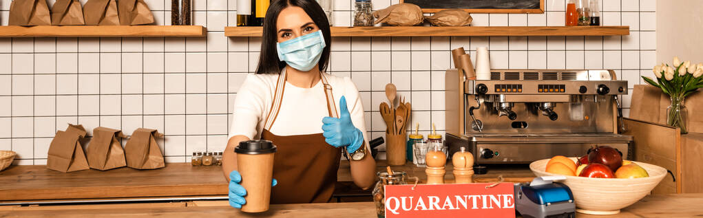 Horizontal image of cafe owner in medical mask showing like sign and paper cup of coffee near table with card with quarantine inscription - Photo, Image
