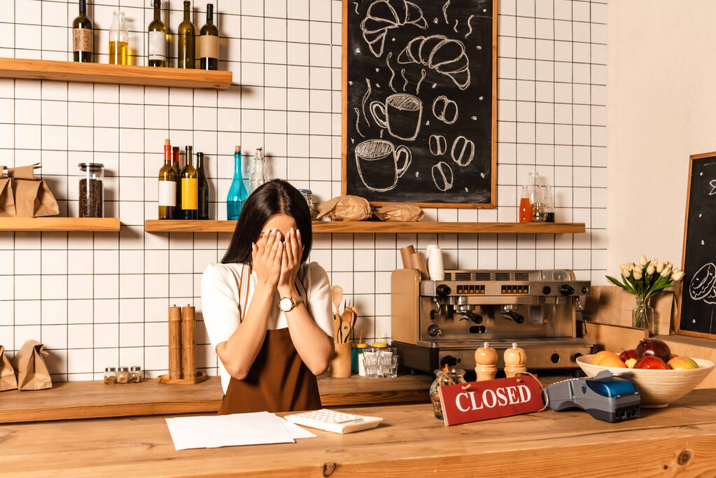 Владелец кафе закрывает лицо у стола бумагами, калькулятором, картой с закрытыми буквами, платежным терминалом и миской с фруктами
  - Фото, изображение