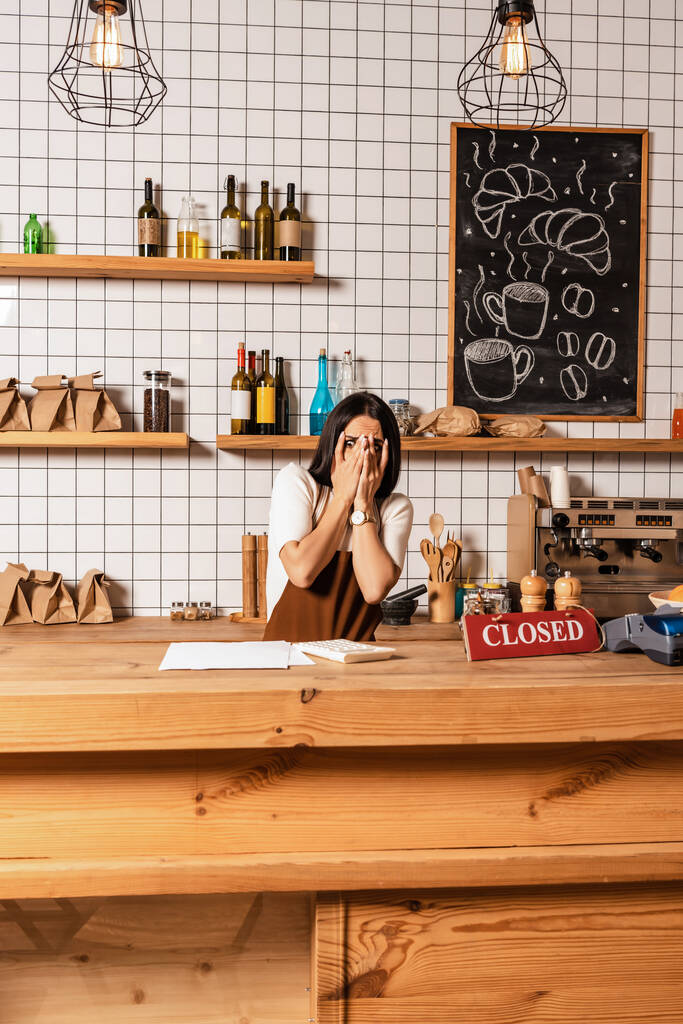 Café proprietário cobrindo rosto perto da mesa com papéis, calculadora, cartão com letras fechadas e terminal de pagamento
 - Foto, Imagem