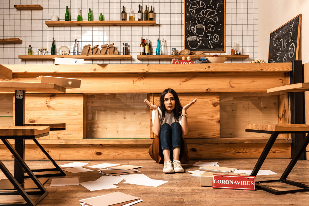 Verwirrter Cafébesitzer blickt auf Kamera am Tisch, Dokumente und Karte mit Coronavirus-Schriftzug auf dem Boden - Foto, Bild