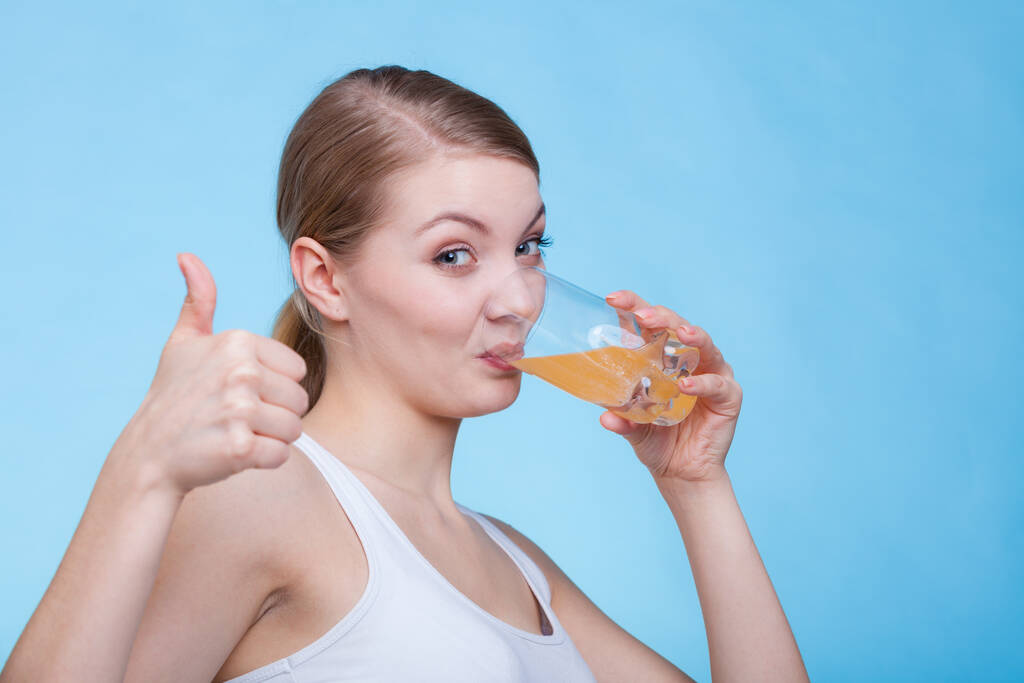 Alimentação, conceito de saúde. Mulher segurando copo de bebida sabor laranja e beber a partir dele mostrando polegar para cima gesto. Estúdio tiro no fundo azul
 - Foto, Imagem