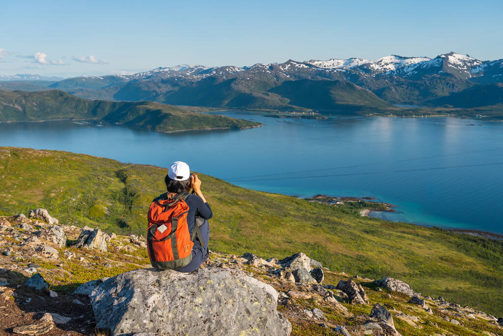 Путешественница с рюкзаком сидит на скале, фотографируя остров Сеня в летний сезон, Норвегия, Скандинавия, Европа
 - Фото, изображение